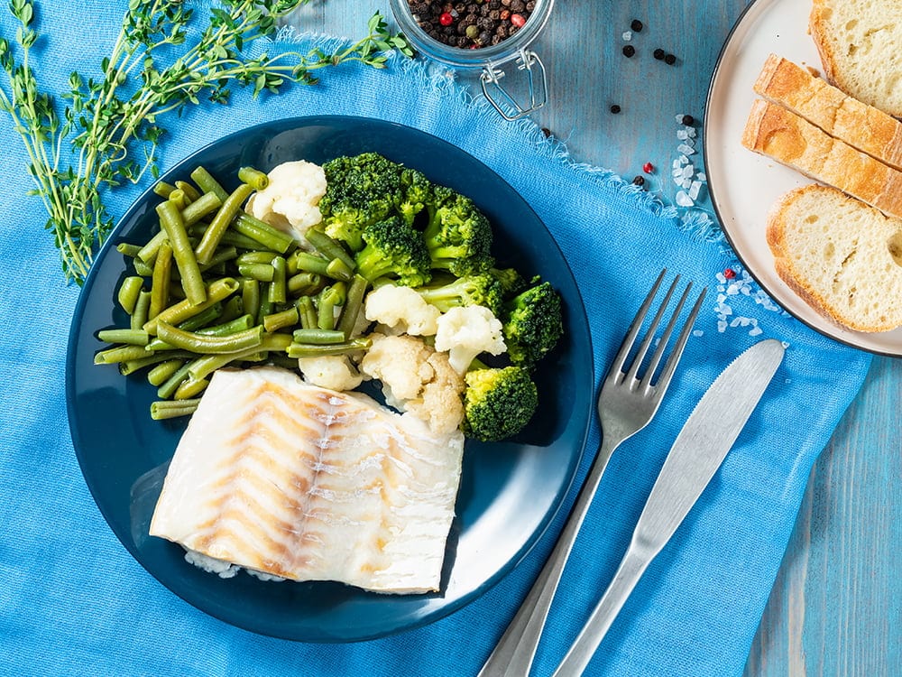 Baked sea fish cod fillet with vegetables – broccoli, green beans and cauliflower  on blue plate, bread, blue napkin, wooden background, top view. Proper diet